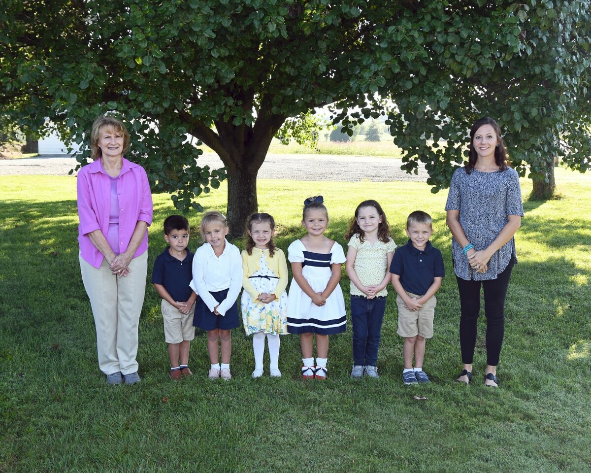 two adult women standing on either side of six preschool age children posing for a class photo